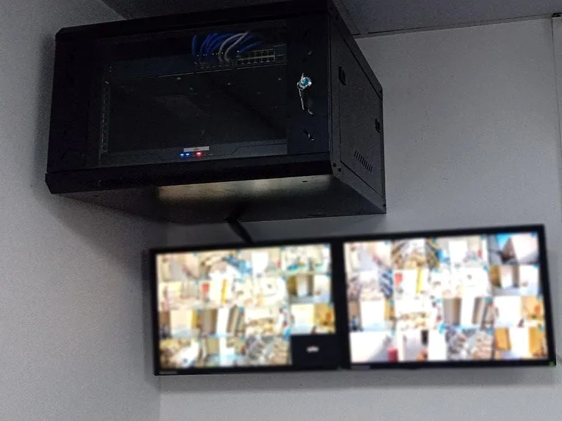 Wall-mounted network cabinet above two monitors showing warehouse CCTV views (screens blurred)